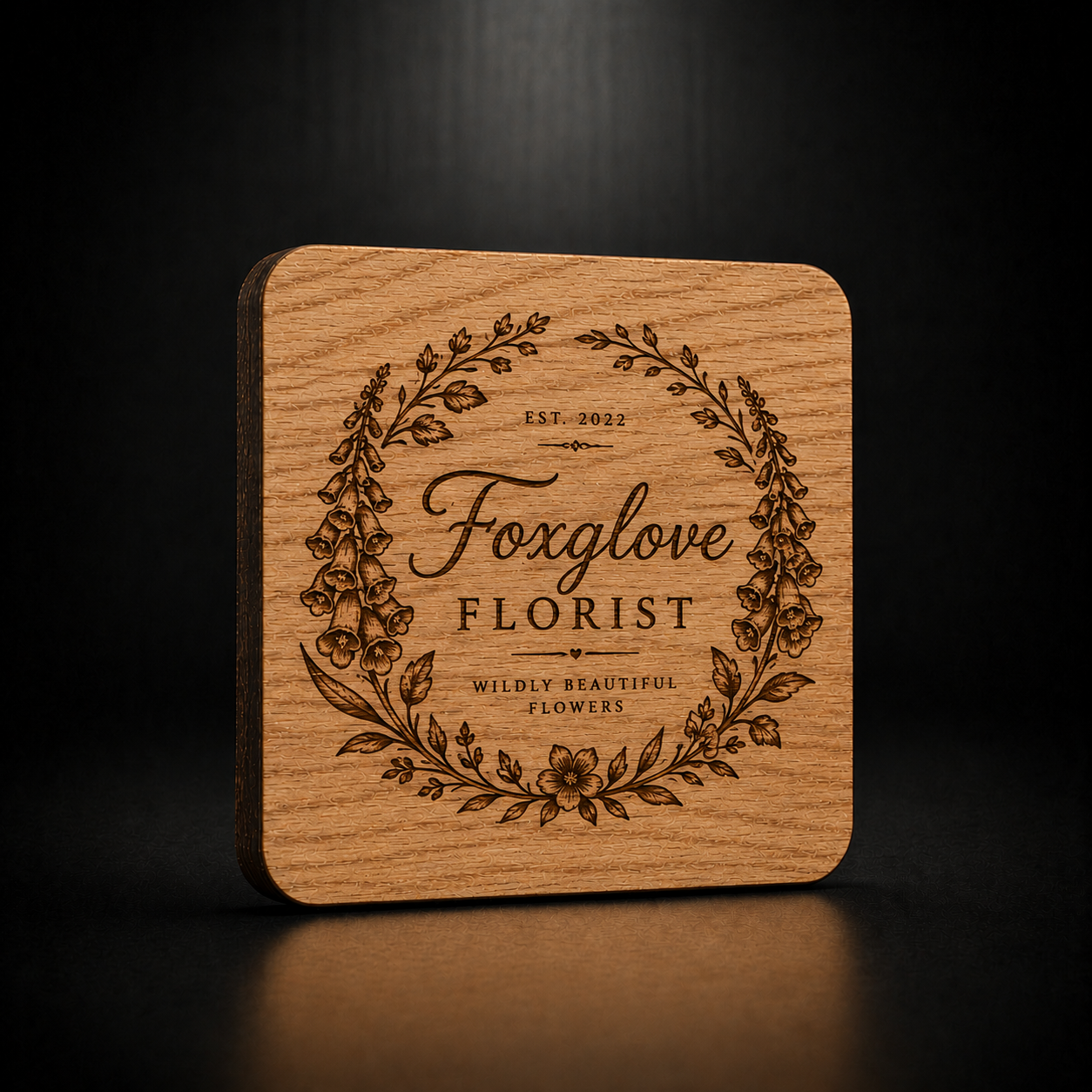A square wooden coaster with rounded corners, featuring a laser-engraved floral wreath and the text 'Foxglove Florist' on a dark, reflective surface.