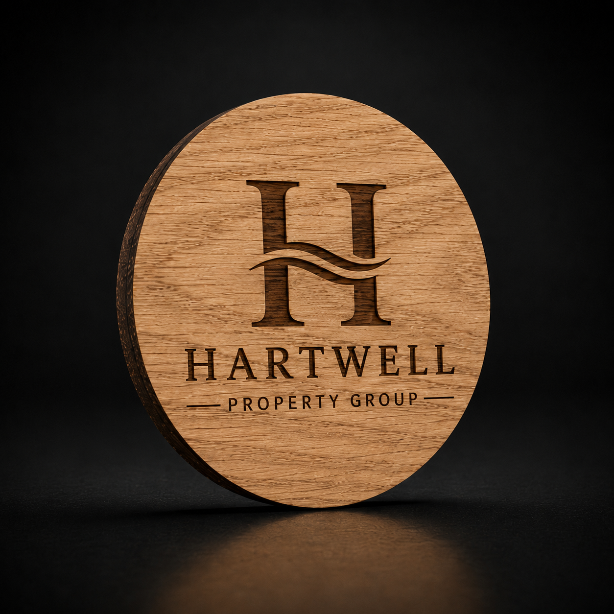 A round wooden plaque or coaster featuring a laser-engraved Hartwell Property Group logo, set against a dark, reflective background.