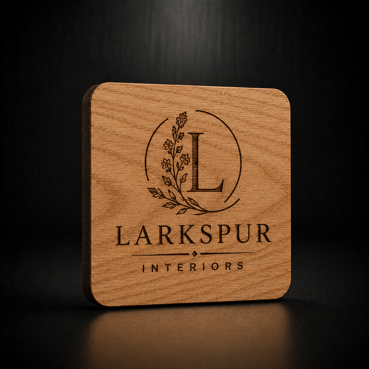 A square wooden coaster with rounded corners, featuring a laser-engraved logo for 'Larkspur Interiors' on a dark wood-grain surface, set against a dark background.