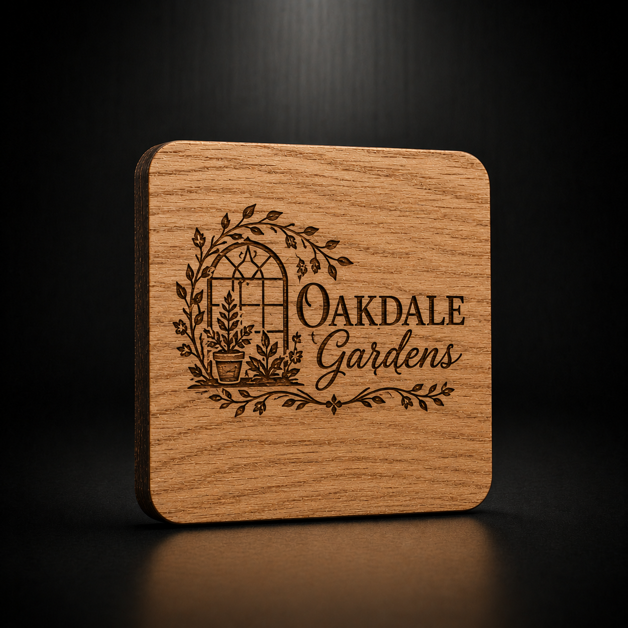 A square wooden coaster with rounded corners, featuring a dark brown engraved logo of an arched window with leaves and the words 'OAKDALE Gardens'.