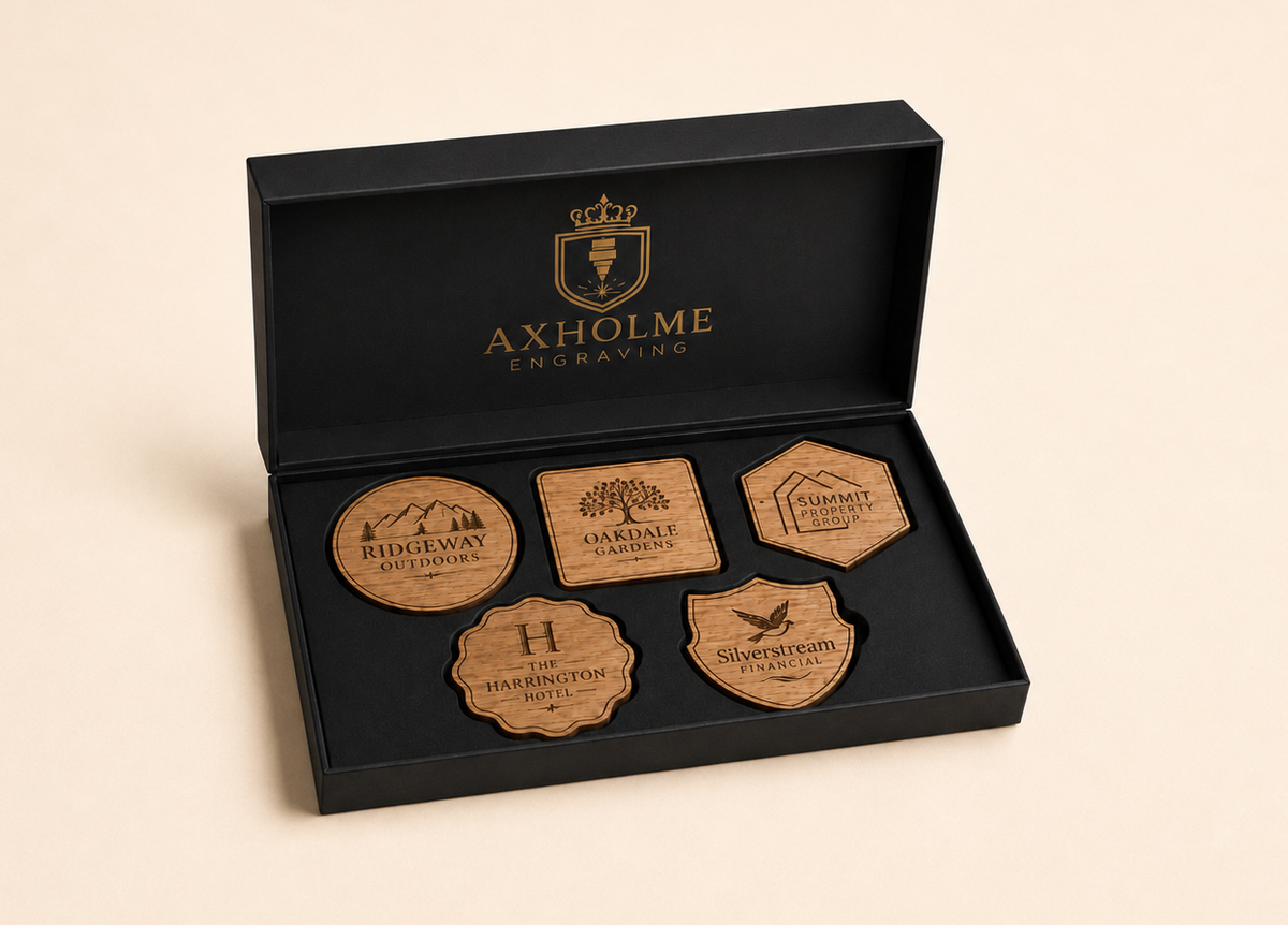 An open black presentation box with gold branding reading AXHOLME ENGRAVING, containing five engraved wooden keepsakes or tags in different shapes, photographed on a warm beige background.