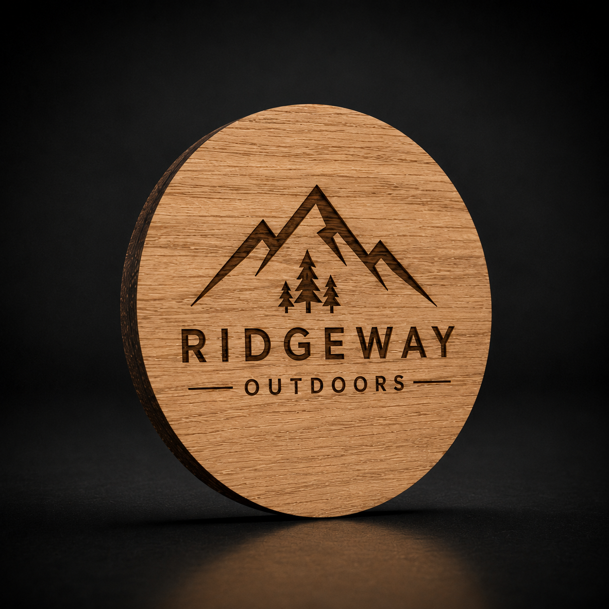 A round wooden coaster with a natural wood grain finish, featuring a dark brown engraved mountain and pine tree logo with the text 'RIDGEWAY OUTDOORS'. The coaster is shown standing upright on a dark, reflective surface against a dark background.