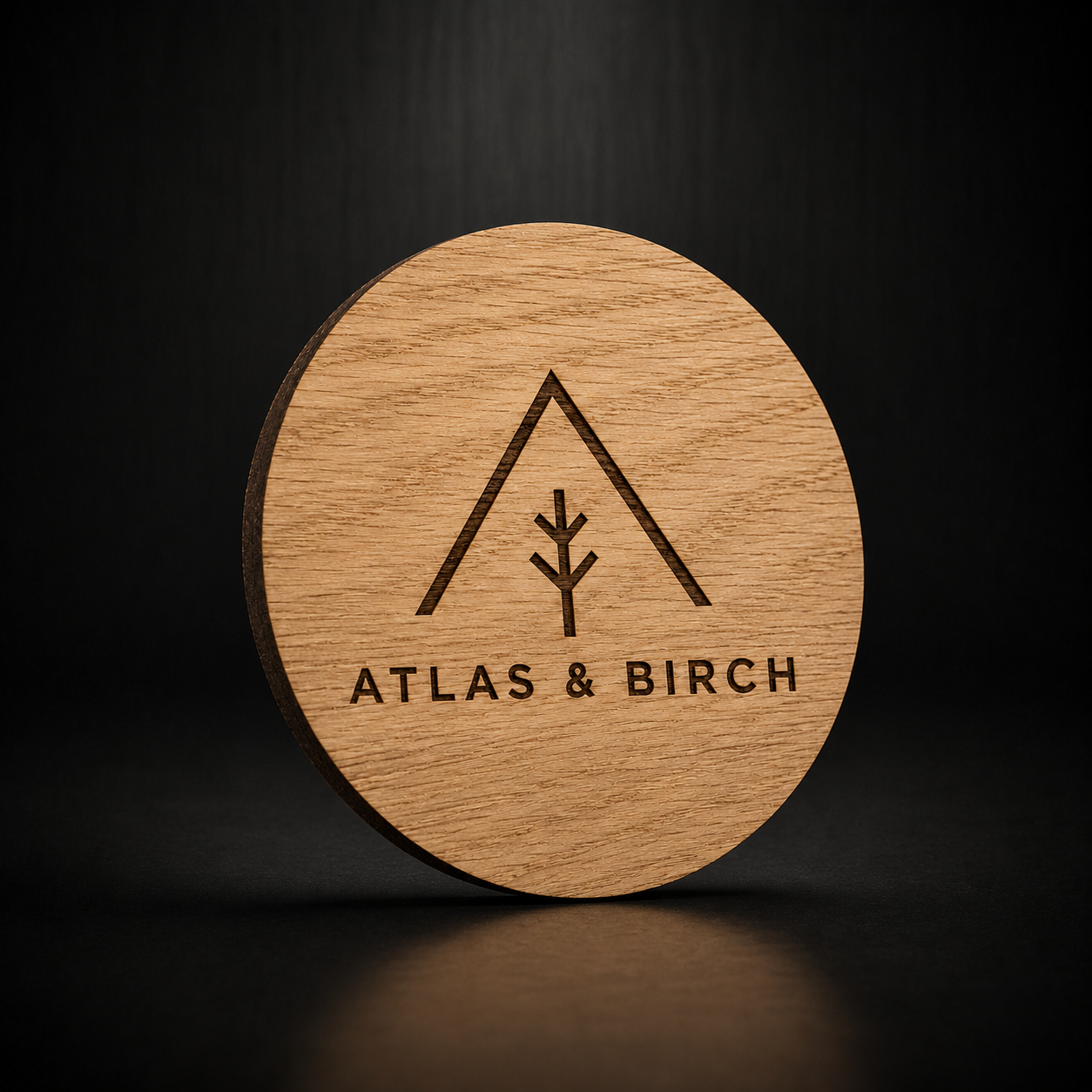 A round natural wood coaster featuring a laser-engraved logo of an upward-pointing tent with a tree inside and the text 'ATLAS &amp; BIRCH' below, set against a dark background.